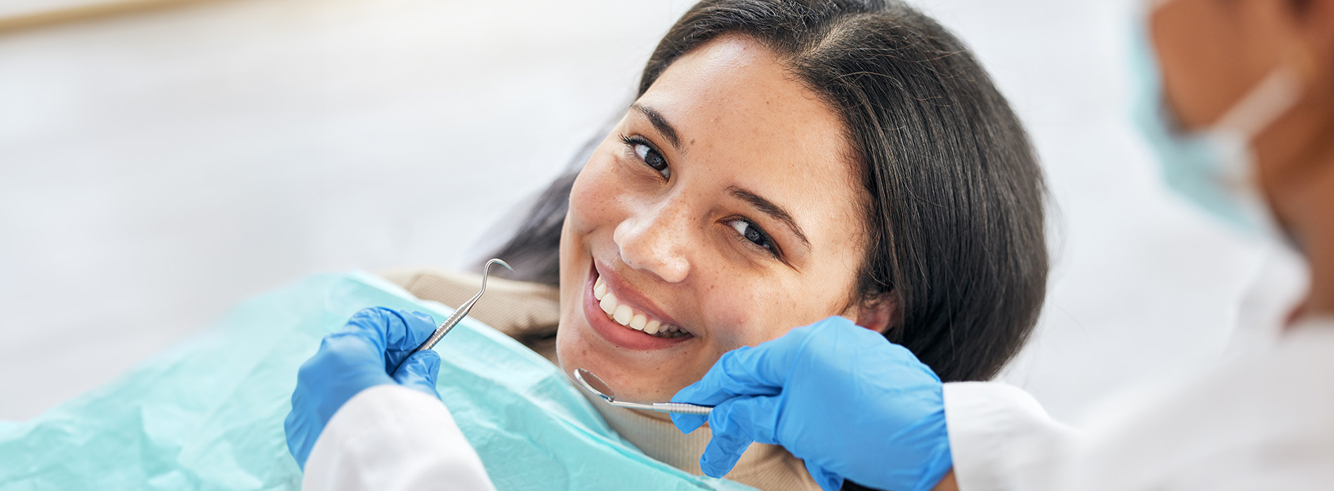 A person is seated in a dental chair, receiving care from a dental professional who stands behind them.